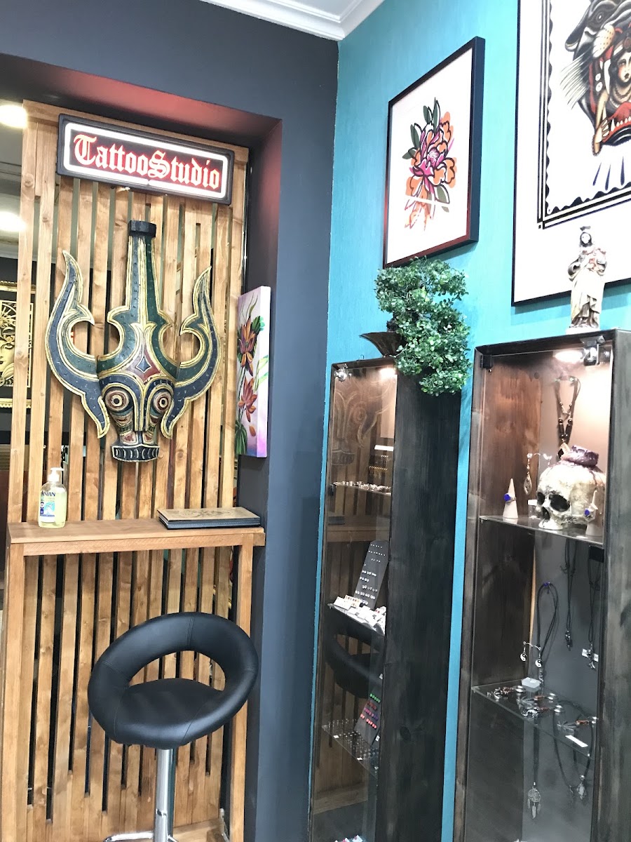 Panel de listones de madera con una máscara tallada y un letrero luminoso rojo que dice TattooStudio; al lado, pared turquesa con láminas enmarcadas y vitrinas de cristal con joyería.