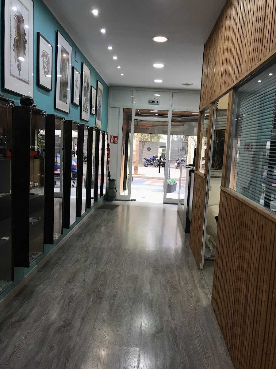 Pasillo hacia la puerta acristalada de la calle: a la izquierda, láminas enmarcadas sobre pared turquesa y vitrinas de joyería; a la derecha, un panel de listones de madera; suelo de laminado gris.