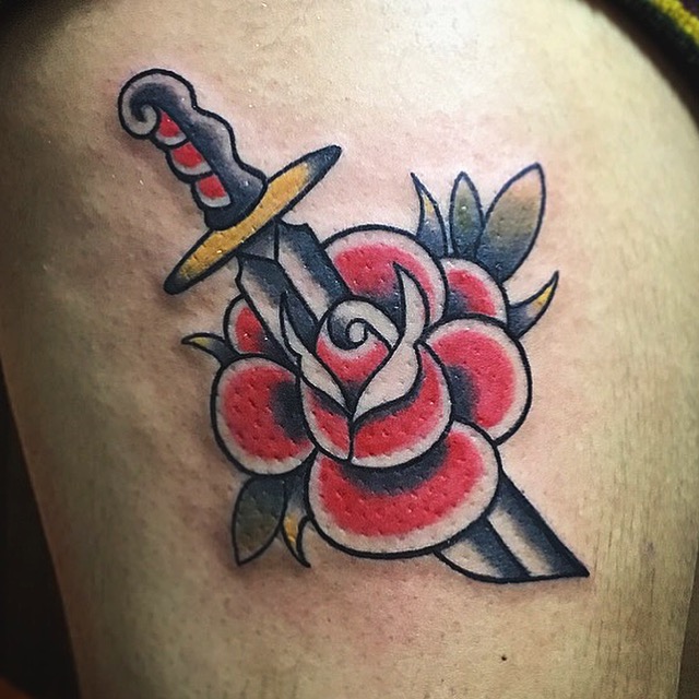 Tatuaje tradicional a color sobre un muslo: una daga que atraviesa una rosa roja con hojas verdes y amarillas.